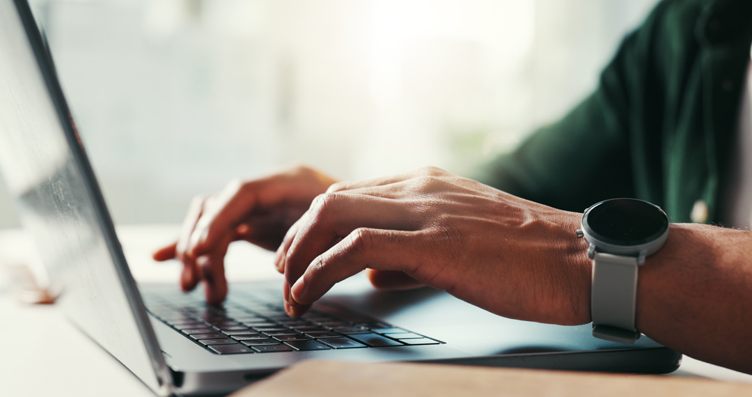 Close-up of a person’s hands wearing a smartwatch typing on a laptop keyboard in soft natural light.